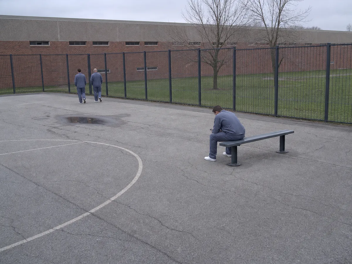 In a fenced recreation yard, a man in a plain slate blue uniform sits sideways on a steel bench looking at a phone in his hands, two others walking the fence line further along the yard.
