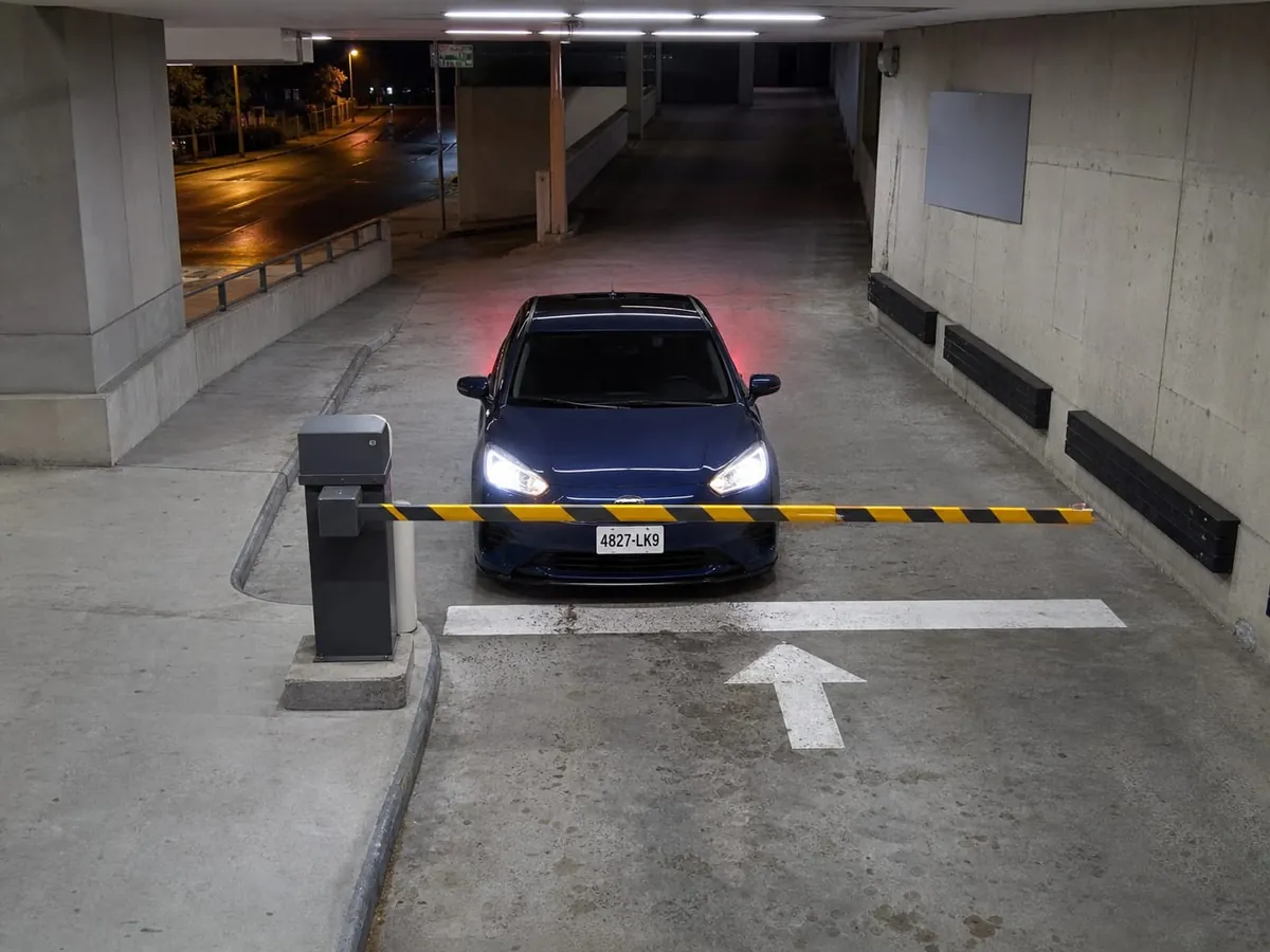 Seen from a camera above a parking garage entrance lane at night, a dark blue sedan waits nose-on at a lowered yellow and black boom barrier with its headlights on, the empty wet street behind it.