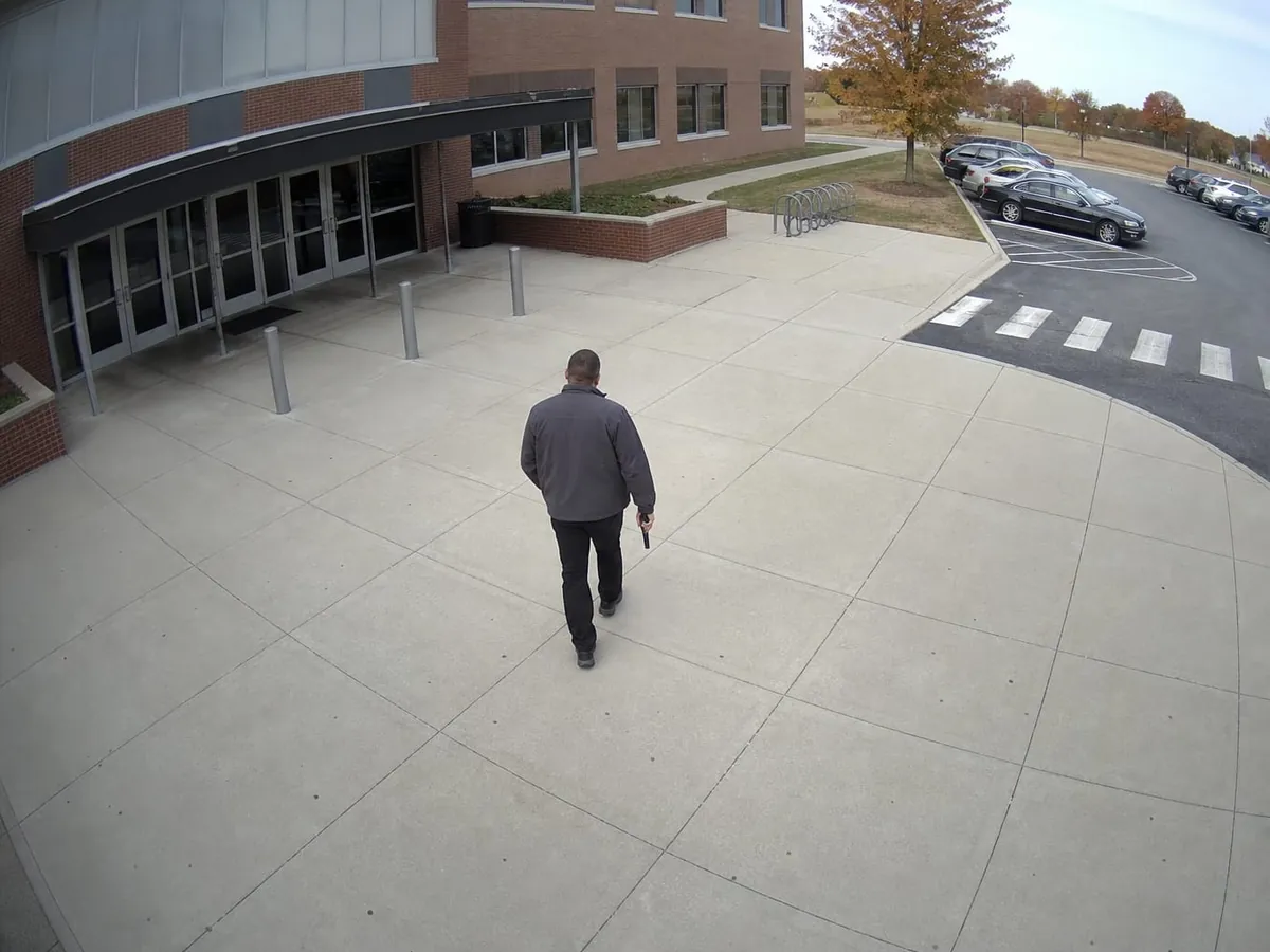 A man in a dark jacket crosses an empty concrete forecourt toward the glazed main doors of a brick school building, seen from a camera above the entrance, a handgun held low against his thigh with the muzzle at the ground.