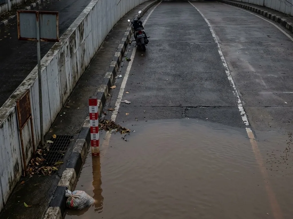 Brown floodwater covers the roadway of a road underpass beside a red and white depth marker post, with a scooter stopped at the water’s edge and debris floating on the surface.