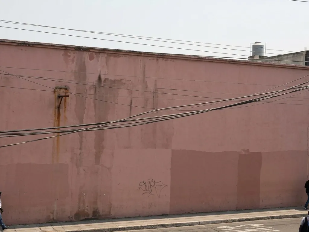 A canted fixed-camera frame filled mostly by a faded rose stucco wall and hazy sky, with utility cables crossing diagonally and only a thin sliver of street and the roofs of two parked vehicles along the bottom edge.