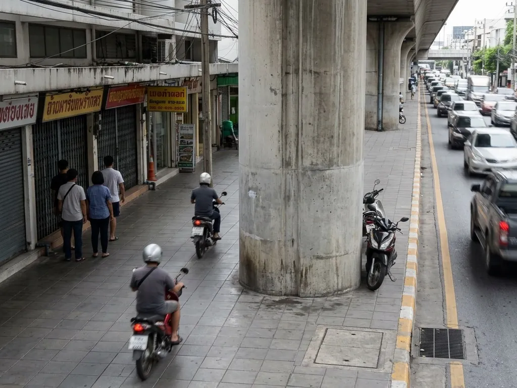 Two small motorbikes ride one behind the other along a Bangkok sidewalk beneath an elevated railway viaduct, weaving around a concrete support pillar, while three pedestrians pressed against the storefronts let them pass.