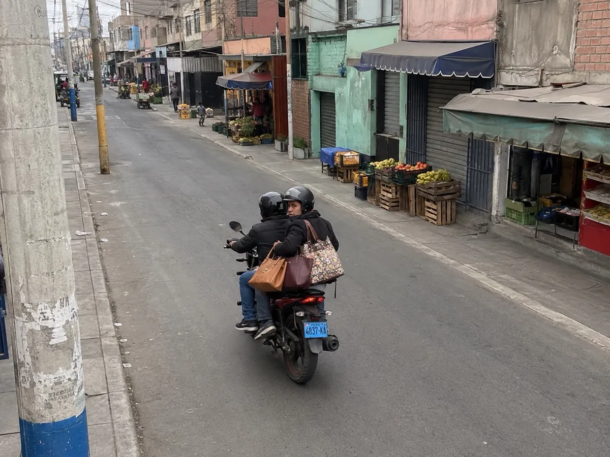 Two helmeted riders on a small motorcycle move away down a Callao market street, the pillion carrying three handbags, the blue rear plate sharp and unobstructed.