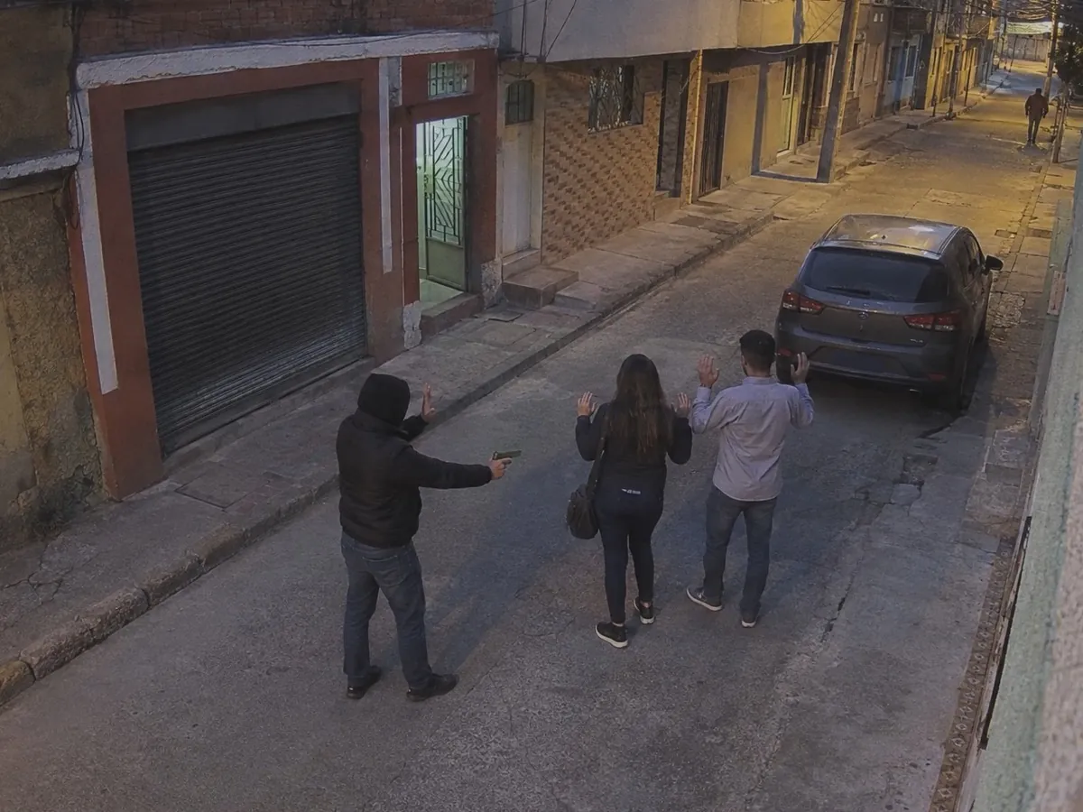 On a Bogotá side street at dusk, seen from a corner-mounted camera, a man in a hooded jacket has just drawn a tan-framed pistol and points it low at a couple whose hands are rising.