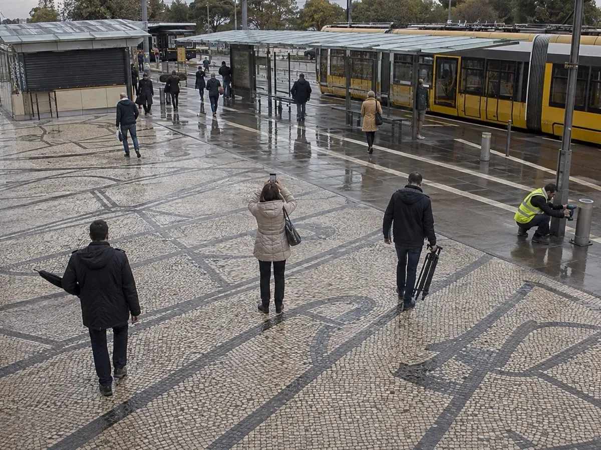 A wide view of a rainy tram interchange forecourt where commuters carry a folded umbrella under an arm, hold a phone up in both hands, carry a tripod by its legs and use a cordless drill at a bollard; nothing in the frame is a firearm.